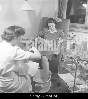 Eine junge Frau in einem Schönheitssalon bekommt eine Pediküre. Der junge Mitarbeiter führt eine Fußpflege durch, bei der die Aufgabe darin besteht, die Füße und Zehennägel zu verschönern. Eine Pediküre hält Ihre Füße in Form und macht sie attraktiver. Während die junge Frau behandelt wird, sitzt sie bequem in einem Sessel und liest eine Zeitschrift. Schweden 1945. Kristoffersson Ref. N108-4 Stockfoto