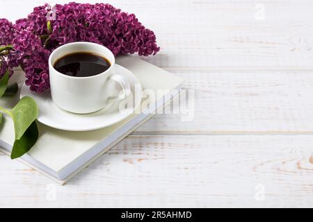 Ein Haufen lila Blumen, ein weißes Buch, eine Tasse schwarzen Kaffee auf einem weißen Holztisch. Leeres Grußkartenmodell. Morgens Lesestunden mit Kaffee Stockfoto