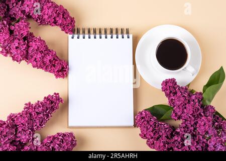 Künstlerische Komposition mit offenem leeren Notizblock, einer Tasse schwarzen Kaffees, lila Blumen auf einem orangefarbenen Tisch. Festliches Desktop-Konzept für das Büro. Morgen-Kaffeetasse Stockfoto