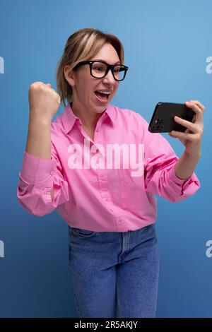 Junge, selbstbewusste blonde Frau mit Pferdeschwanz und Brille in einem trendigen rosa Hemd für das Büro, um Videos anzusehen Stockfoto