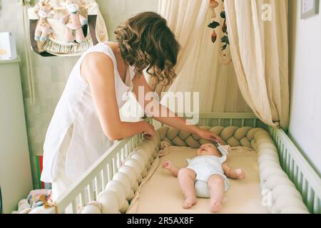 Junge Mutter kümmert sich um ihr kleines Baby, Kind liegt in einem Kinderbett Stockfoto