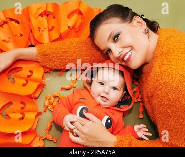 Halloween-Porträt einer glücklichen jungen Mutter und eines süßen Babys mit orangefarbener Kürbis-Jacke Stockfoto