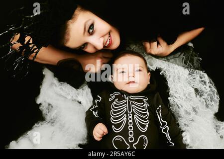 Halloween-Porträt eines niedlichen Babys, das Skelettkostüm trägt, auf schwarzem Hintergrund mit Spinnennetz posiert, Mutter trägt Hexenkostüm und liegt n Stockfoto