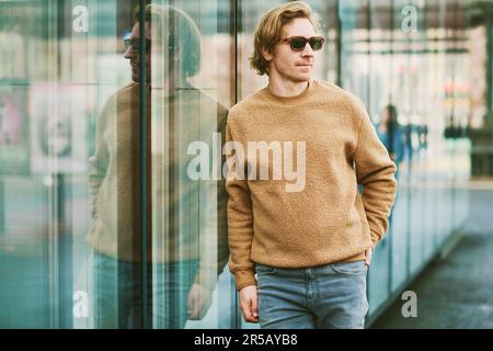 Außenporträt eines gutaussehenden jungen Mannes mit Sonnenbrille und beigefarbenem, flauschigem Fleece-Pullover, der sich neben einer Spiegelwand im Straßenhintergrund der Stadt posiert Stockfoto