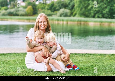 Außenporträt einer glücklichen jungen Mutter mit zwei reizenden Kindern, die an einem schönen Sommertag am See oder Fluss spielen Stockfoto