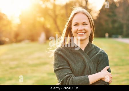 Romantisches Außenporträt einer hübschen jungen Frau im Frühlingspark bei Sonnenuntergang, die sich in einer grünen Jacke bedeckt Stockfoto
