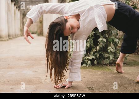 Ein weiblicher Yoga in wildem Ding posiert auf einer traumhaften grünen Terrasse eines Landschaftsgartens mit Säulen in schwarz-weiß fließendem, lässigem Outfit. Barefoo Stockfoto