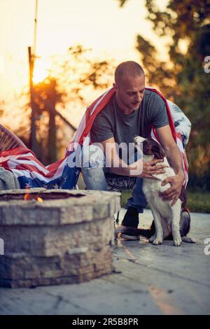 Junger Mann streichelt Border Collie Hündchen, genießt Zeit im Freien, mit der US-Flagge auf seinem Rücken, sitzt am Kamin in einem gemütlichen Stuhl. Unabhängigkeit Da Stockfoto