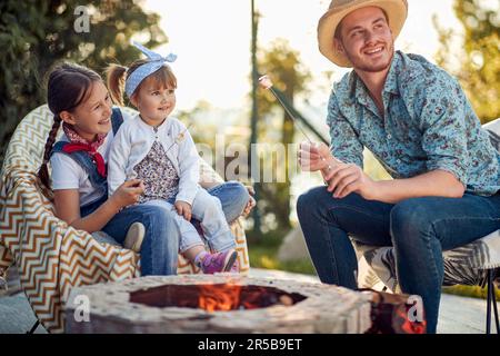 Fröhlicher Vater, der tagsüber an einem sonnigen Sommertag mit seinen zwei kleinen Töchtern in einem gemütlichen Landhaus im Freien Marshmallows röstet. Lebensstil, Zusammensein Stockfoto