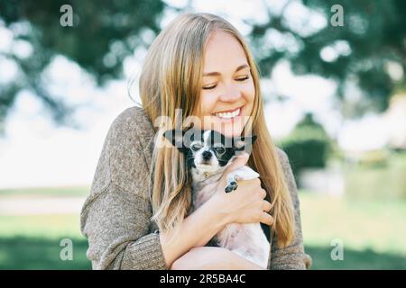 Freiluftporträt einer glücklichen jungen Frau, die einen kleinen chihuahua schwarz-weißen Hund umarmt Stockfoto