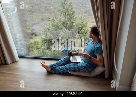 Junger Mann in lässiger Kleidung und barfuß sitzt in der Nähe eines Panoramafensters mit Laptop. Männlicher Freiberufler arbeitet am Computer und trinkt Tee oder Kaffee. Wunderbar Stockfoto