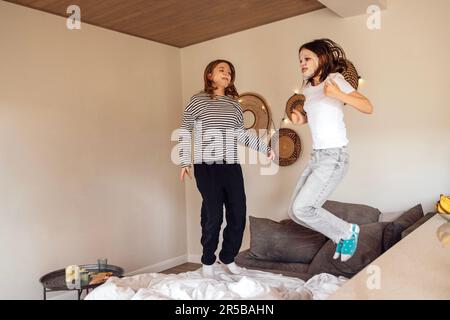 Süße Teenager-Mädchen mit Einweg-Stoff feuchtigkeitsspendenden Schönheitsmasken auf ihren Gesichtern springen auf das Sofa. Reizende weibliche Freunde in legerer Kleidung haben Stockfoto