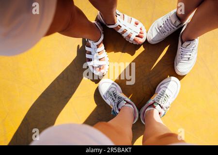 Drei Paar Familienfüße stehen zusammen auf gelbem Hintergrund. Süßes Selfie von drei Paaren, weibliche Beine in weißen Sandalen und Turnschuhen. Freundschaft Stockfoto