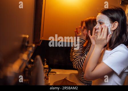 Zwei süße Teenager Mädchen tragen Einweg-Stoff feuchtigkeitsspendende Schönheitsmasken auf ihren Gesichtern im Badezimmer. Süße weibliche Freunde machen Spa-Behandlungen zu Hause. Stockfoto