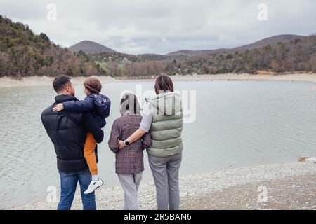 Glückliche Familie mit zwei Kindern am See. Die Leute tragen warme Jacken und Mäntel. Mom, Dad, Teenager und Vorschultochter, die sich zurückhalten. Mann, Frau, Stockfoto