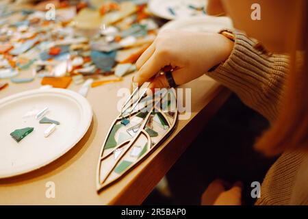 Weibliche Hand mit Ringen, die während der Mosaikherstellung ein Glasstück mit einer Pinzette festhält. Teenager-Mädchen stecken ein grünes Stück Glas in die Form eines Baumblattes. Mehrspaltig Stockfoto