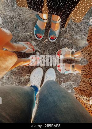 Vier Paar Füße stehen zusammen auf trockenem Gras. Süßes Selfie mit vier Paaren, weiblichen Beinen mit roter Pediküre in weißen Sandalen und Turnschuhen. Freundschaft Stockfoto