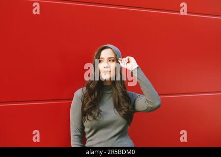 Außenporträt einer jungen hübschen Frau, die auf rotem Hintergrund posiert und Khaki-Hoodie trägt Stockfoto