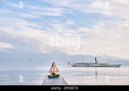 Genfer Seenlandschaft, Frau, die Yoga am Dock praktiziert, Dampfboot schwimmt im Hintergrund Stockfoto