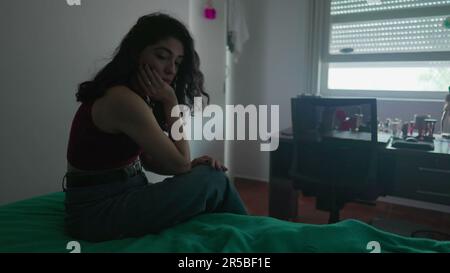Eine nachdenkliche junge Frau sitzt am Bett mit nachdenklichem Ausdruck. Weibliche Person in 30s mit inneren Emotionen Stockfoto