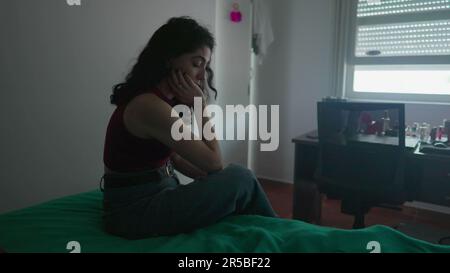 Eine nachdenkliche junge Frau sitzt am Bett mit nachdenklichem Ausdruck. Weibliche Person in 30s mit inneren Emotionen Stockfoto