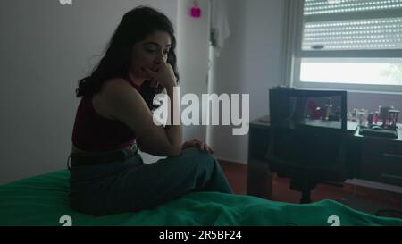 Eine nachdenkliche junge Frau sitzt am Bett mit nachdenklichem Ausdruck. Weibliche Person in 30s mit inneren Emotionen Stockfoto