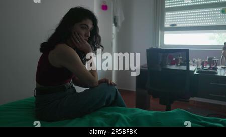 Eine nachdenkliche junge Frau sitzt am Bett mit nachdenklichem Ausdruck. Weibliche Person in 30s mit inneren Emotionen Stockfoto