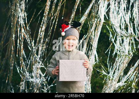 Außenporträt eines fröhlichen und aufgeregten kleinen Jungen, der neben einem dekorierten Kiefernbaum posiert, eine Geschenkbox in der Hand hält, Rentierohren trägt und einen Weihnachtsmann-Hut trägt Stockfoto