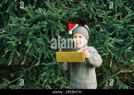 Außenporträt eines fröhlichen und aufgeregten kleinen Jungen, der neben einer Kiefer posiert, eine Geschenkbox in der Hand hält, Rentierohren trägt und einen Weihnachtsmann-Hut trägt Stockfoto