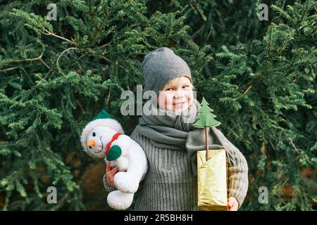 Außenporträt eines fröhlichen, süßen Jungen, der neben einer Kiefer posiert, eine Geschenkbox und ein Schneemannspielzeug in der Hand hält, einen Wurmschal und einen Hut trägt Stockfoto