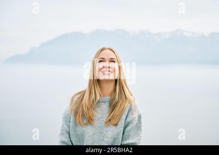 Außenporträt einer glücklichen, wunderschönen jungen Frau, die sich in den Bergen über den Wolken entspannt und einen grauen Pullover trägt Stockfoto