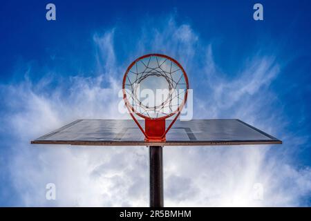 Basketballkorb von unten gesehen mit dem blauen Himmel mit einigen Wolken, dem orangefarbenen Korb und dem weißen Rückenbrett Stockfoto