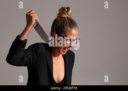 Bedrohliche blonde Frau mit angesammelten Haaren, schwarzer Jacke und Brille trägt ein großes Messer in ihren Händen Stockfoto