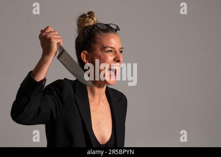 Bedrohliche blonde Frau mit angesammelten Haaren, schwarzer Jacke und Brille trägt ein großes Messer in ihren Händen Stockfoto