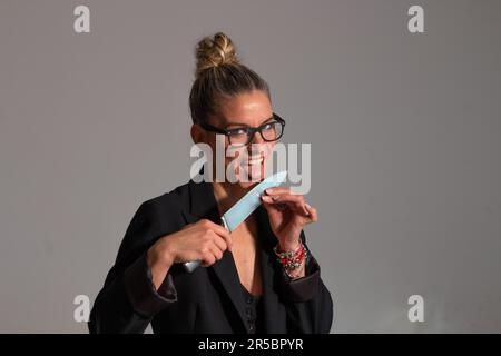 Bedrohliche blonde Frau mit angesammelten Haaren, schwarzer Jacke und Brille trägt ein großes Messer in ihren Händen Stockfoto