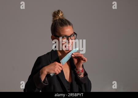 Bedrohliche blonde Frau mit angesammelten Haaren, schwarzer Jacke und Brille trägt ein großes Messer in ihren Händen Stockfoto