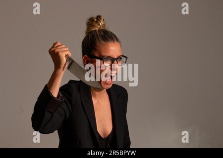 Bedrohliche blonde Frau mit angesammelten Haaren, schwarzer Jacke und Brille trägt ein großes Messer in ihren Händen Stockfoto