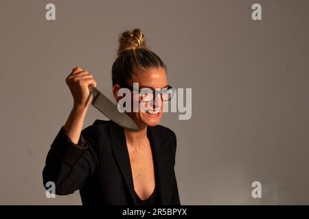 Bedrohliche blonde Frau mit angesammelten Haaren, schwarzer Jacke und Brille trägt ein großes Messer in ihren Händen Stockfoto