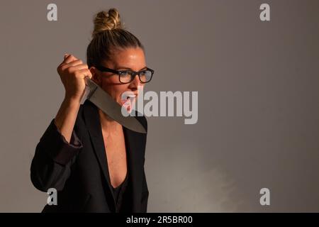 Bedrohliche blonde Frau mit angesammelten Haaren, schwarzer Jacke und Brille trägt ein großes Messer in ihren Händen Stockfoto