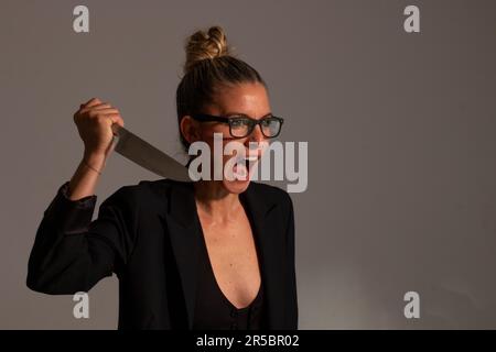 Bedrohliche blonde Frau mit angesammelten Haaren, schwarzer Jacke und Brille trägt ein großes Messer in ihren Händen Stockfoto