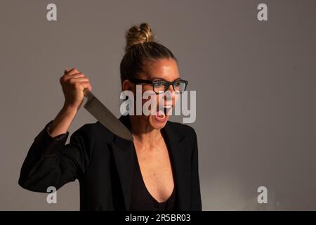 Bedrohliche blonde Frau mit angesammelten Haaren, schwarzer Jacke und Brille trägt ein großes Messer in ihren Händen Stockfoto