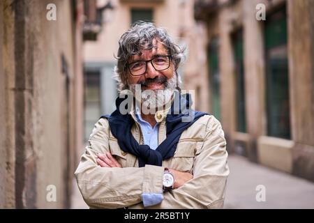 Ein grauhaariger Mann kreuzte die Arme und lächelte fröhlich, als er in die Kamera sah und auf der Straße stand. Stockfoto