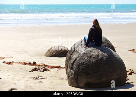 Eine Frau, die auf Moeraki Felsen in Neuseeland sitzt und die Landschaft des Ozeans genießt Stockfoto