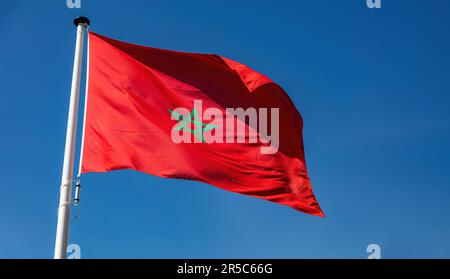 Marokko-Zeichen-Symbol. Marokkanische Staatsflagge auf Flaggenmast, winkend im Wind, blauer Hintergrund Stockfoto