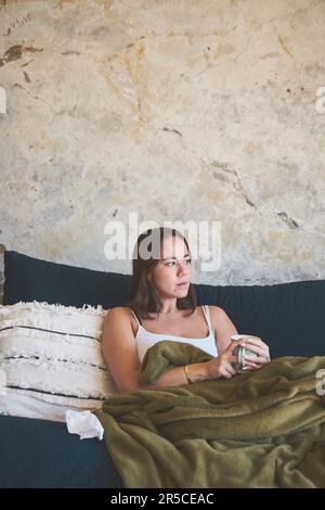 Die Quarantäne hatte ihre eigenen Herausforderungen. Eine junge Frau, die sich zu Hause auf dem Sofa von einer Krankheit erholt und ein warmes Getränk trinkt. Stockfoto