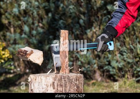 Hacken von Holz durch Spalten der Axt mit fliegendem Holzstamm auf grünem, verschwommenem Waldhintergrund. Nahaufnahme des menschlichen Arms mit scharfem Axt in der Hand mit Arbeitshandschuh. Stockfoto