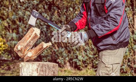 Arbeitender Holzschneider hält Spaltaxt in der Hand und hackt Holzstämme auf Holzblock. Brennholzaufbereitung mit einem modernen Stahlaxialwerkzeug. Energiekrise. Stockfoto