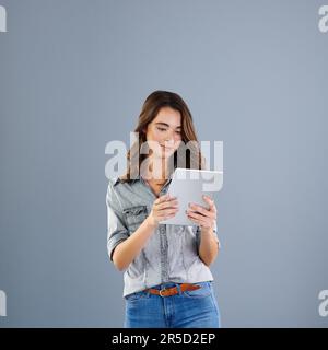 Sie ist immer verbunden. Studioaufnahme einer attraktiven jungen Frau, die ihr digitales Tablet vor grauem Hintergrund verwendet. Stockfoto