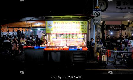 Street Food-Stand auf dem Huaxi Street Tourist Night Market in Taipei, Taiwan; Tische und Restaurantgäste sitzen im Hintergrund. Stockfoto