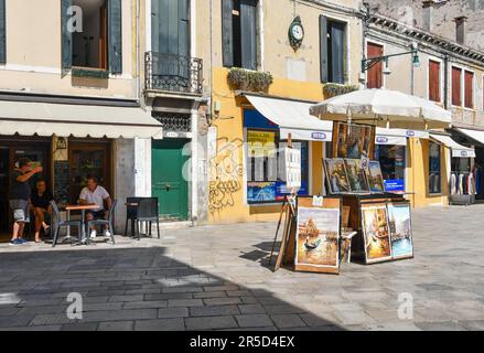 Souvenirgemälde unter einem Schirm in Rio Terà San Leonardo mit Leuten im Straßencafè im Sommer, Venedig, Veneto, Italien Stockfoto